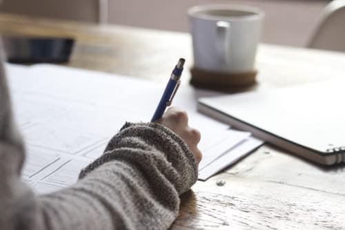 person-writing-on-brown-wooden-table-near-white-ceramic-mug